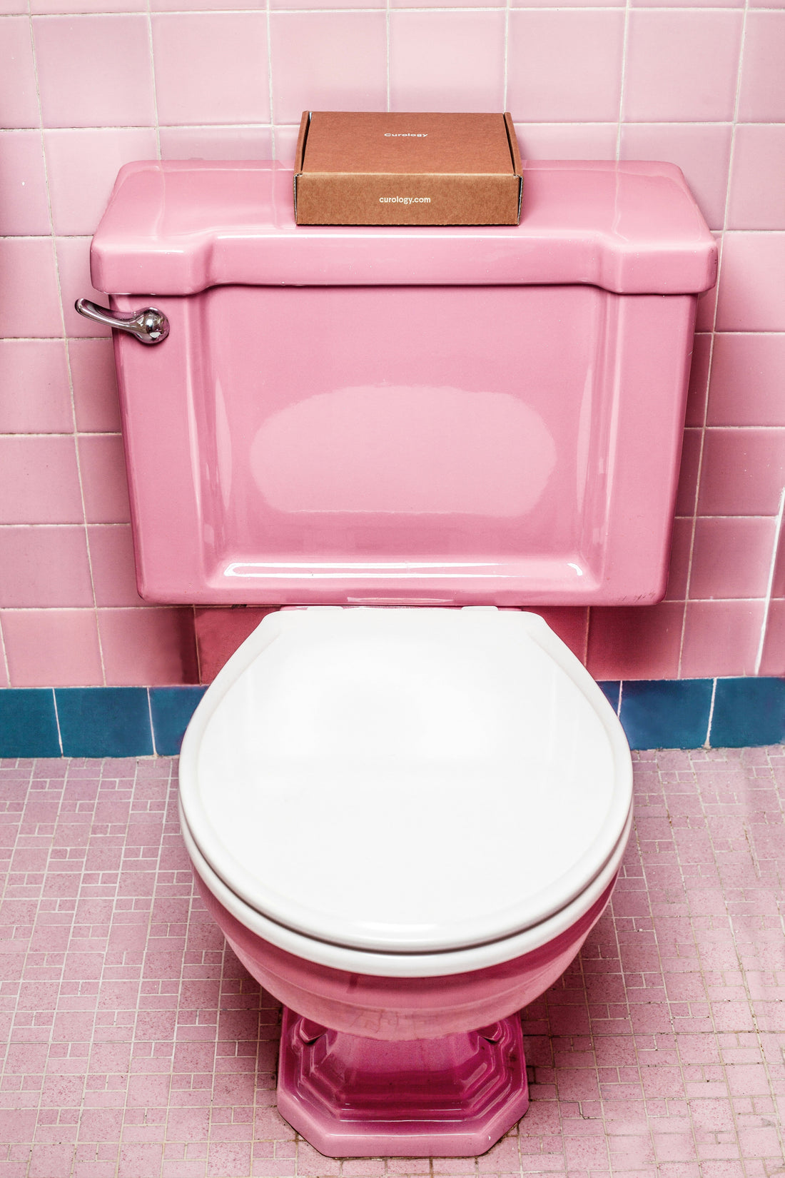 Roze toilet met witte bril in een badkamer met roze tegels. Op de spoelbak staat een klein bruin doosje.