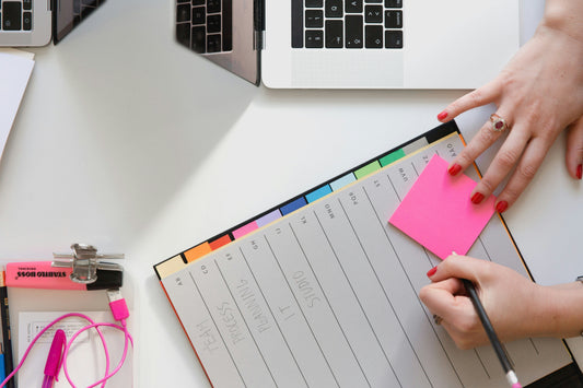 Een wit bureau met daarop een planner, en handen met rode nagellak die op een roze post-it gaan schrijven.